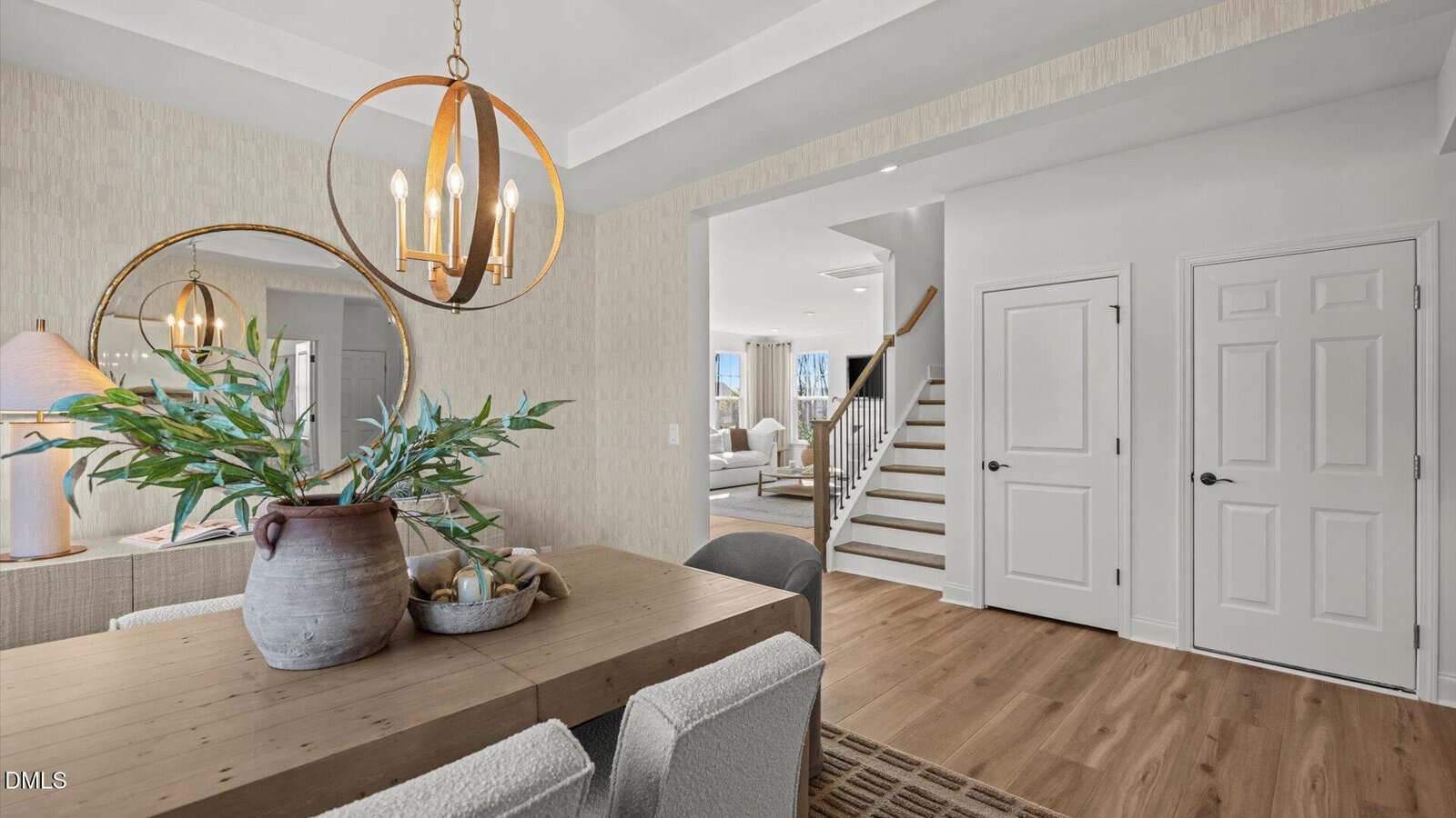 Dining area with a wooden table, pottery vase, mirror, and light fixture. View into a living room, staircase, and two white doors on the right, all with light wood flooring and neutral decor.