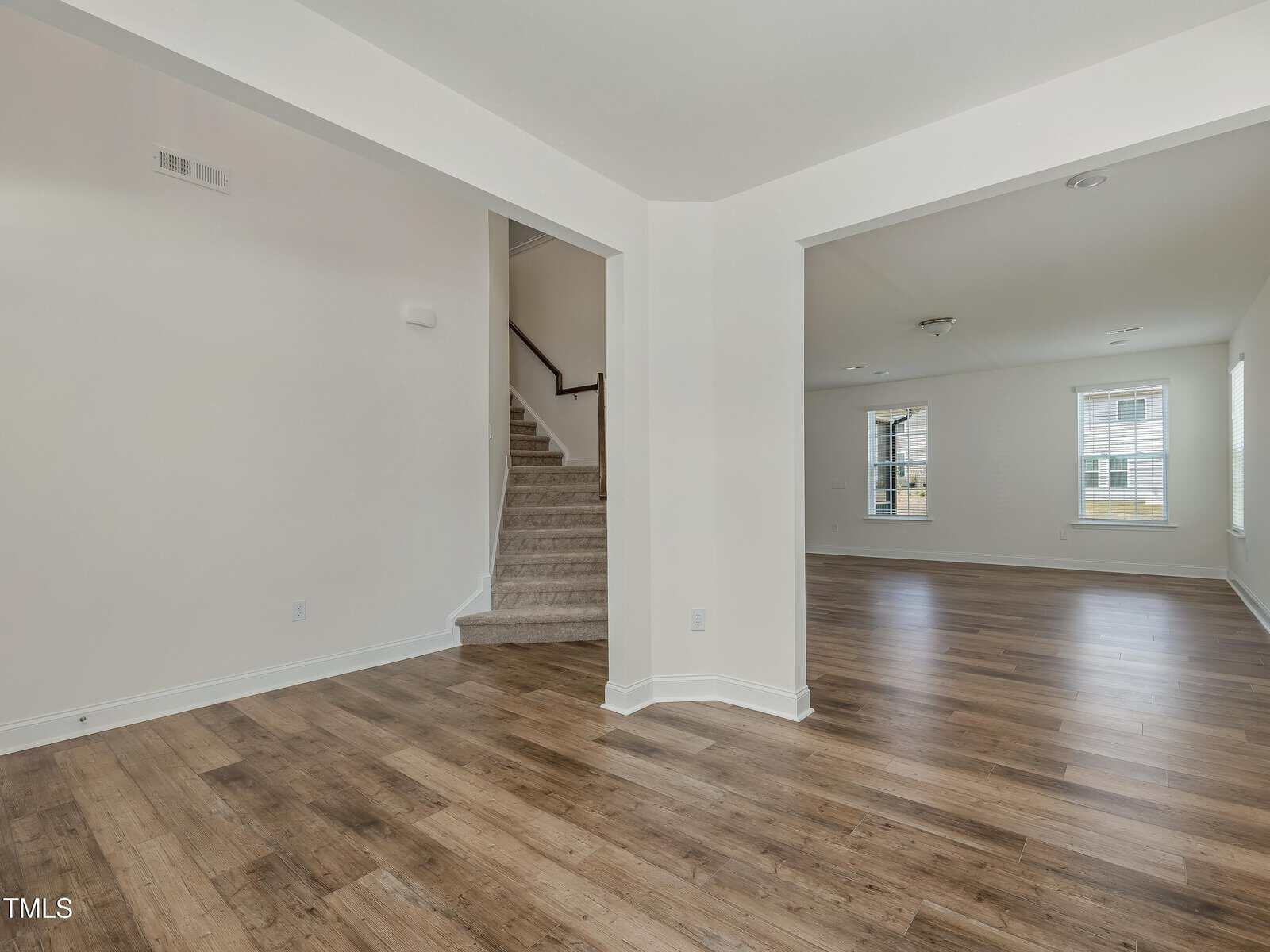 Open space interior with wood flooring, white walls, a staircase with carpeted steps leading up, and multiple large windows letting in natural light. Ceiling lights are visible in both rooms.