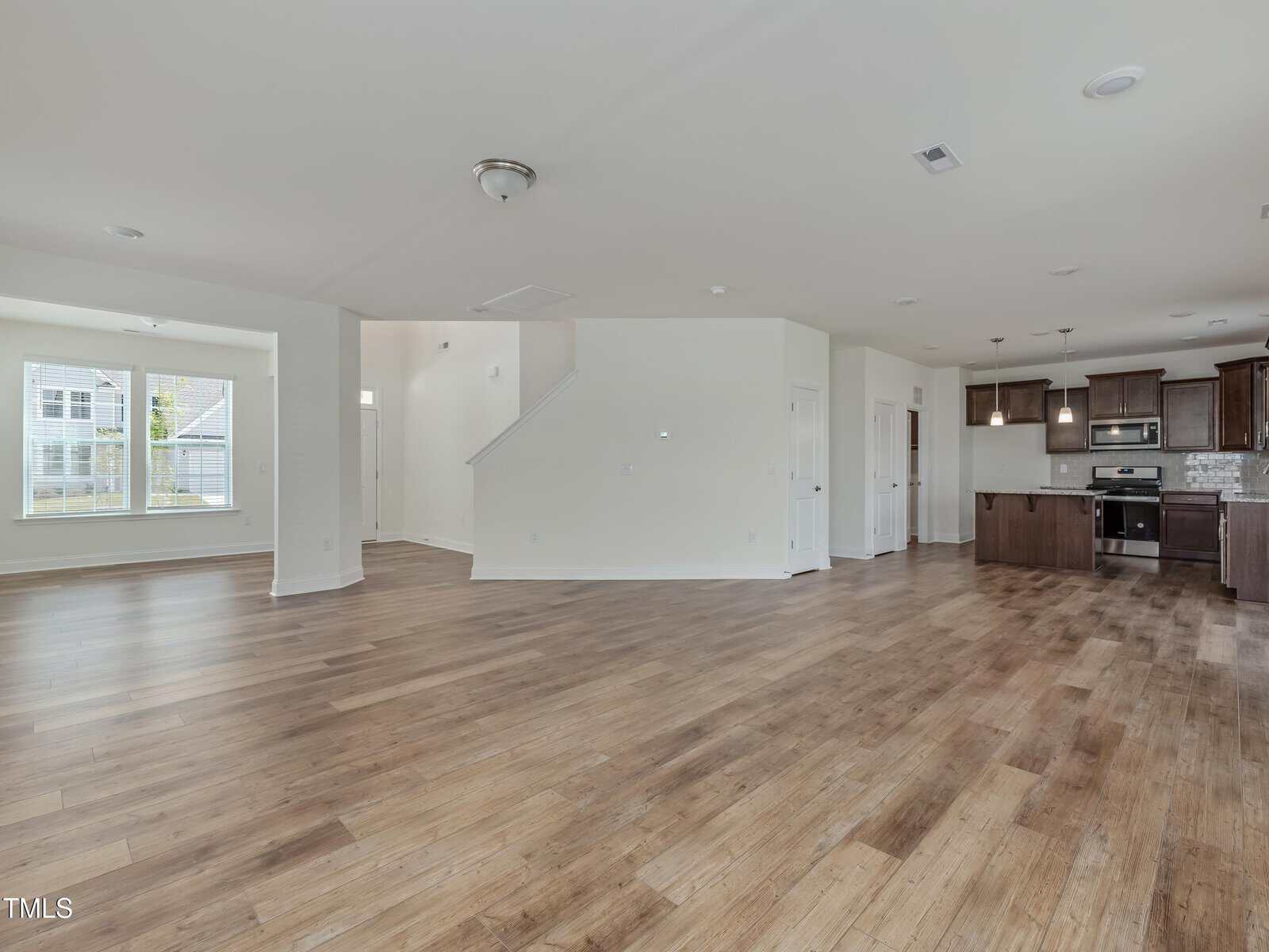 Open-concept living area with wood floors, neutral walls, large windows, staircase, and a modern kitchen featuring dark cabinets, stainless steel appliances, and pendant lighting. The space is unfurnished and well-lit.