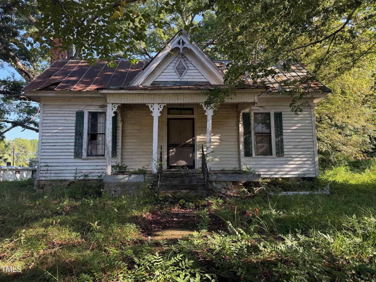 A small, weathered white house at 4079 Buckhorn Road in Bullock, with peeling paint, a rusty metal roof, overgrown vegetation, and a front porch with decorative trim. Two shuttered windows flank a closed central door as trees cast shadows over the scene.
