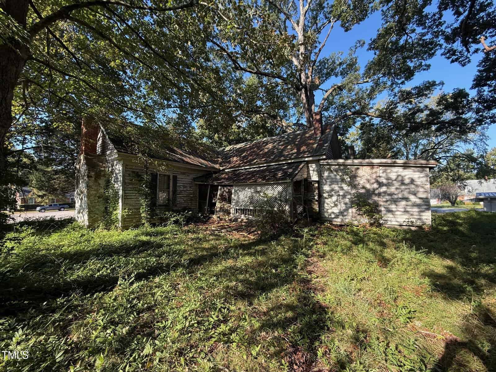 A small, weathered house with peeling white paint sits in the shade of large trees. The yard is overgrown with grass and weeds, and sunlight filters through the branches, casting shadows on the building.