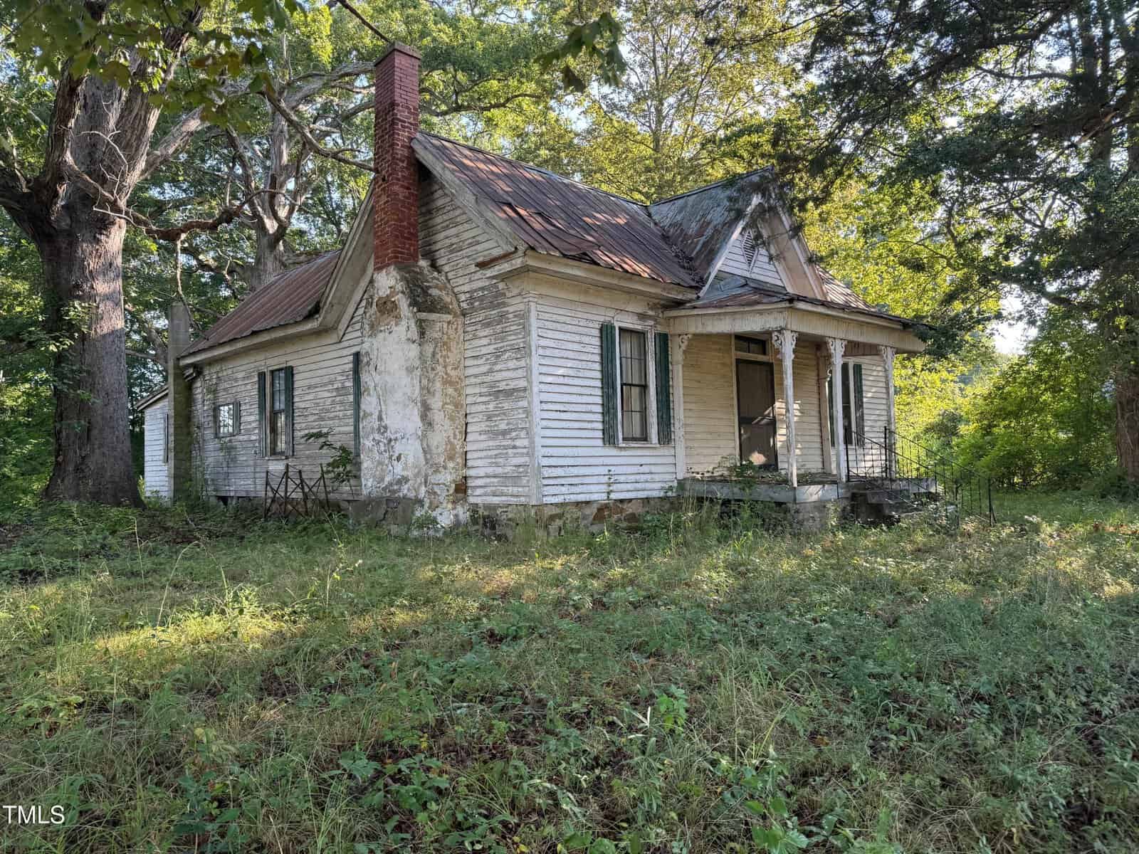 An old, weathered white house with peeling paint, a rusted metal roof, and a brick chimney sits in an overgrown yard surrounded by tall trees and grass.