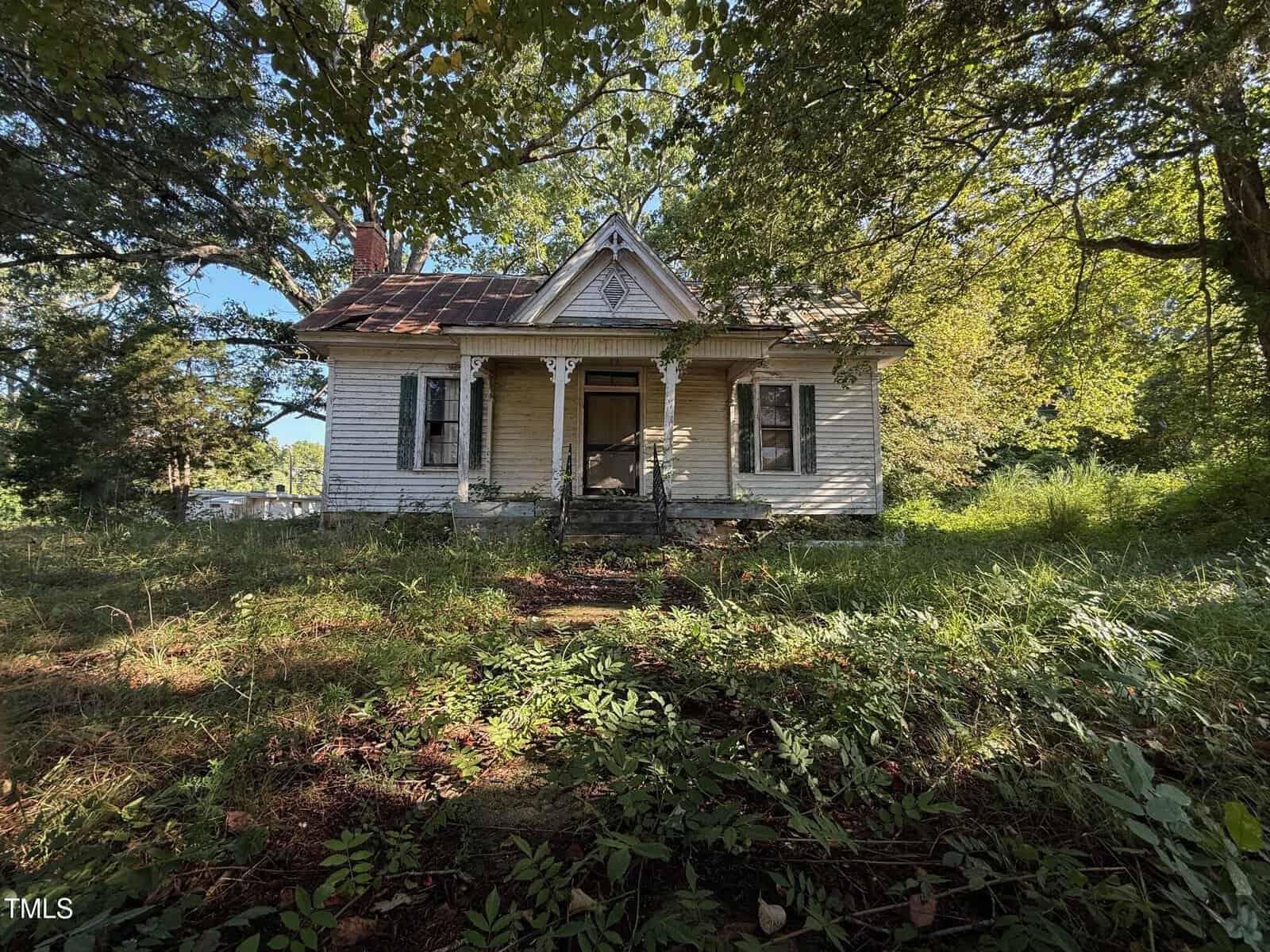 An old, white, wooden house with a covered front porch, overgrown grass, and dense greenery surrounding the property. The house appears weathered, and sunlight filters through the trees above.