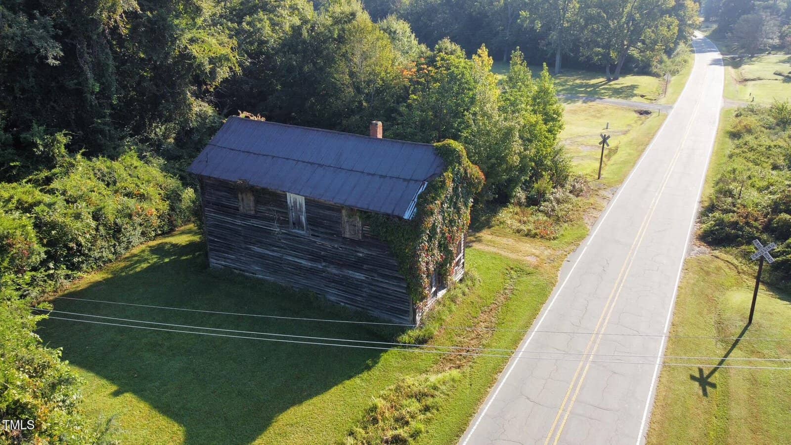 A weathered, abandoned wooden building with a metal roof sits beside a rural road. Ivy climbs parts of the structure. Green grass and trees surround the area under daylight. Power lines run across the foreground.