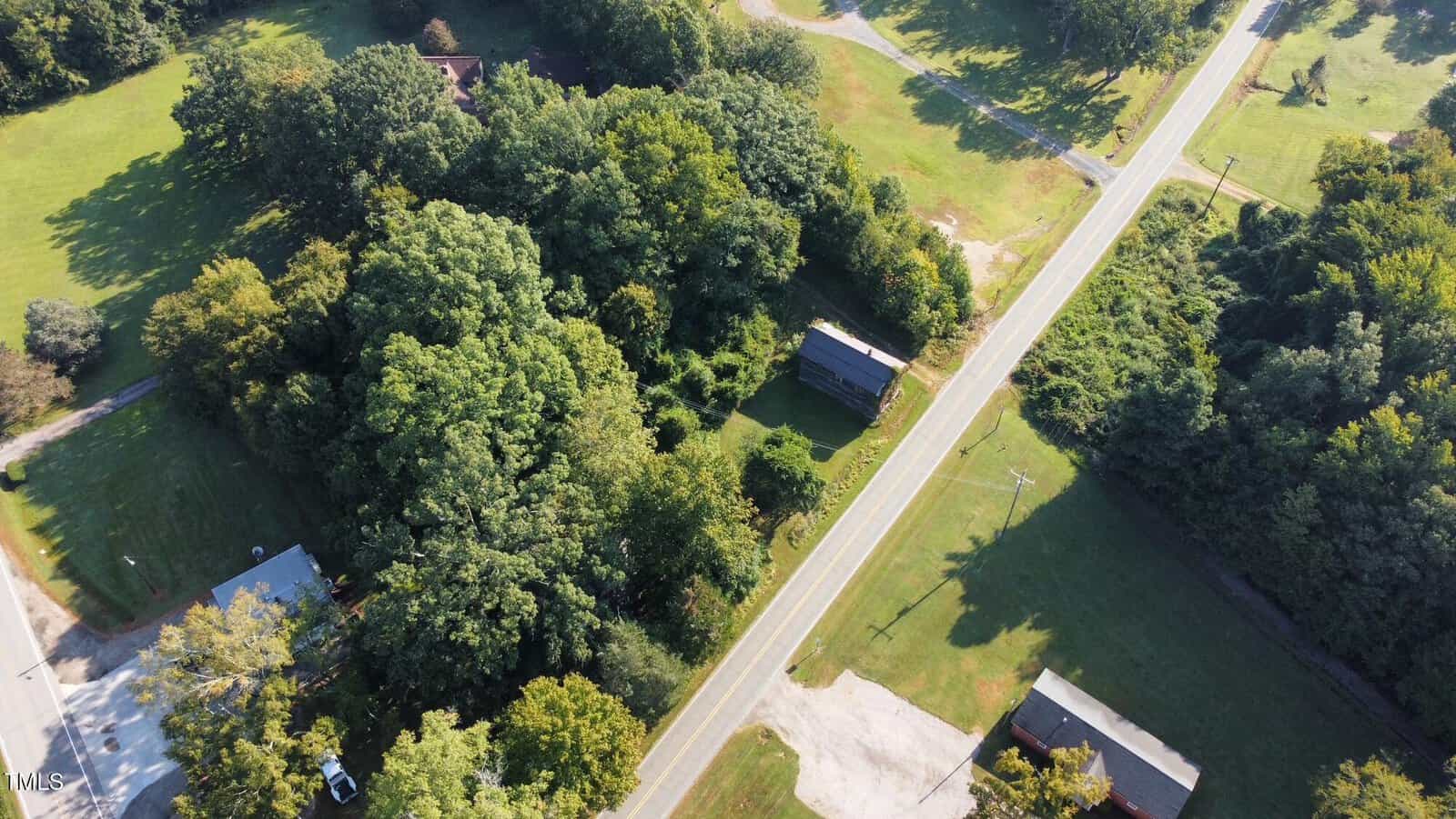 Aerial view of a rural area with a house surrounded by dense trees near a road, nearby buildings, and grassy lawns. The road runs vertically through the right side of the image.