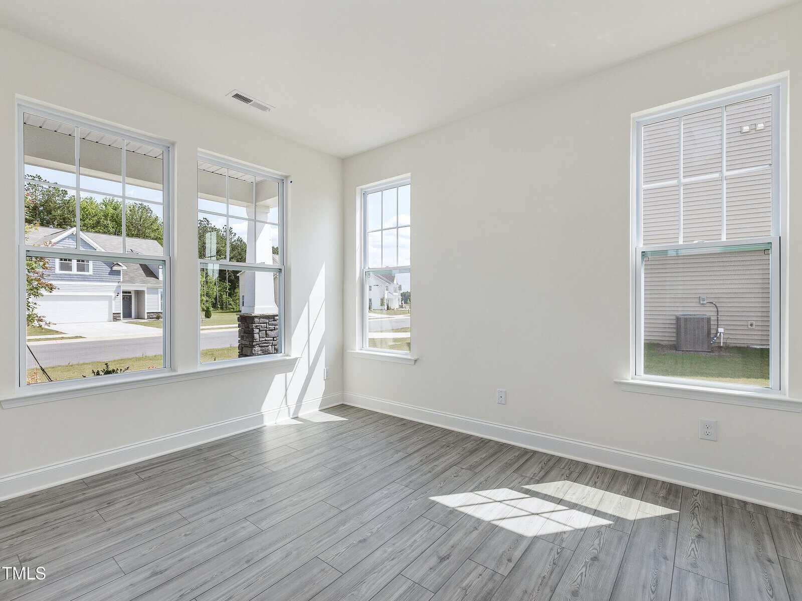 Bright, empty room with light gray wood flooring, white walls, and three large windows letting in natural light. The view outside shows neighboring houses and a street. The room appears clean and modern.