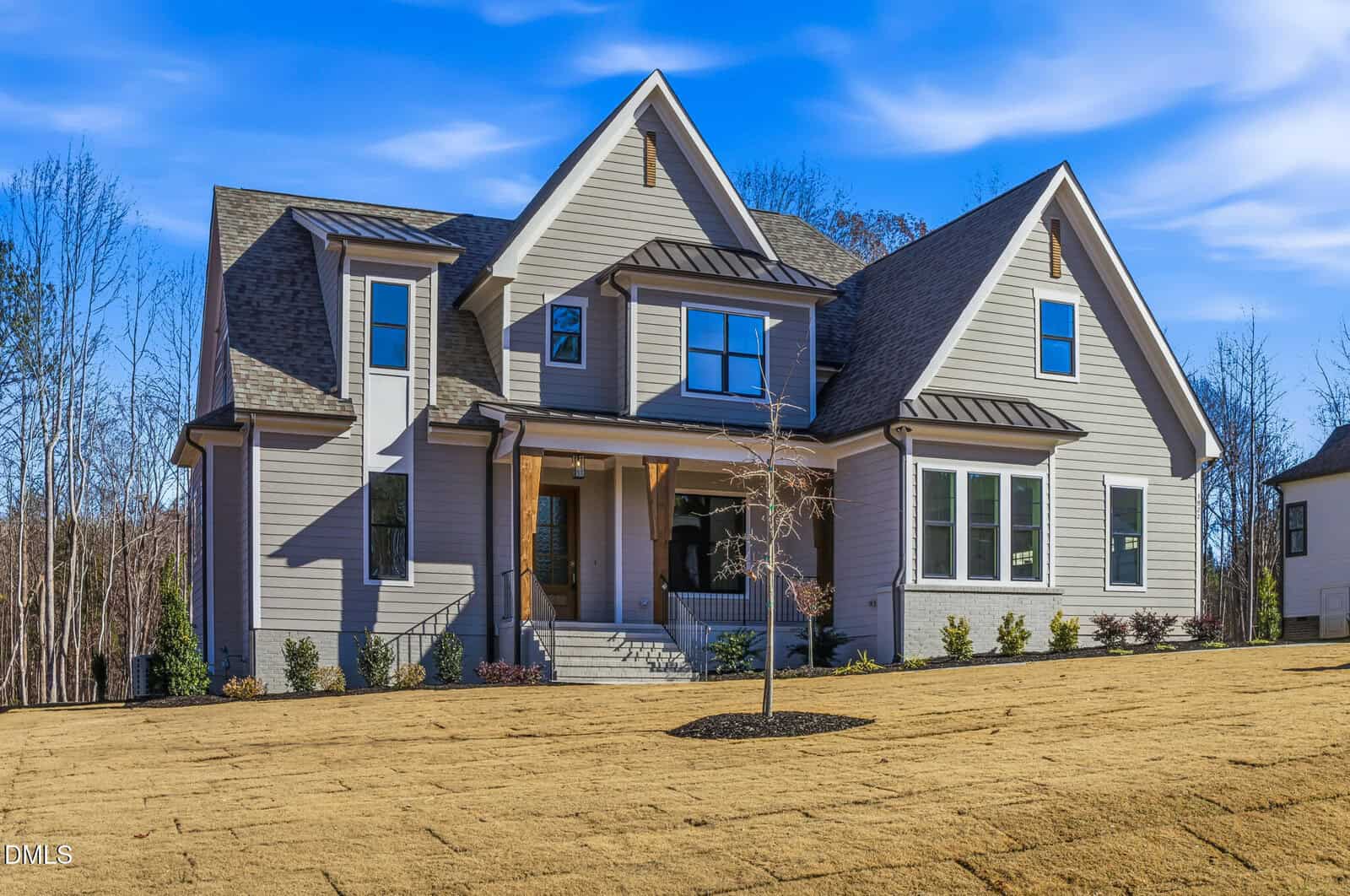 Two-story modern house with gray siding, large front porch, and multiple gables; new landscaping with a small tree and freshly laid sod in the front yard, under a clear blue sky.