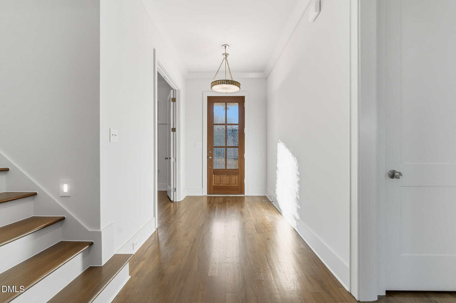 A bright hallway with wooden floors, white walls, a staircase on the left, a wooden front door with glass panes, and a modern chandelier hanging from the ceiling. Two closed doors are visible on the right side.