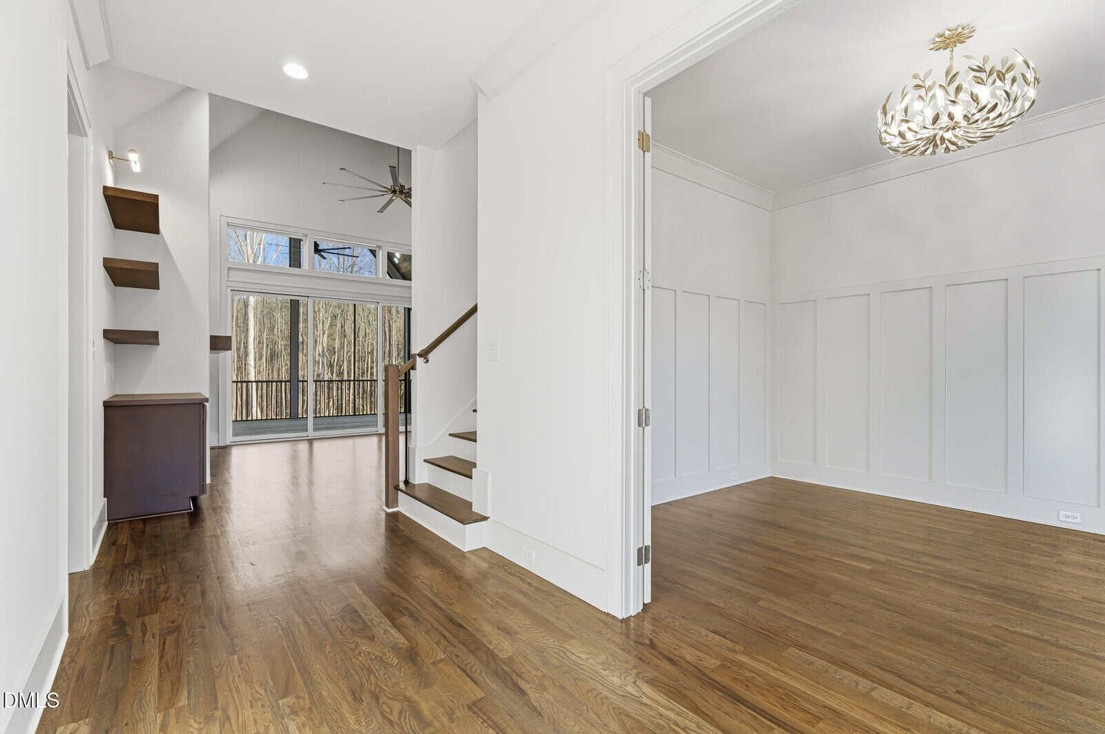 A modern, unfurnished interior with hardwood floors, white walls, a staircase, and a separate room with decorative wall paneling. Large windows and glass doors in the background provide natural light and views of trees.