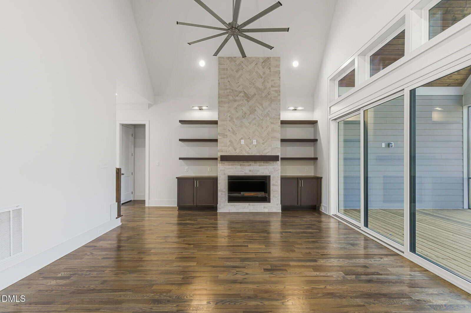 A modern, empty living room with hardwood floors, a stone fireplace with built-in shelves on both sides, a large ceiling fan, and sliding glass doors leading to an enclosed porch.