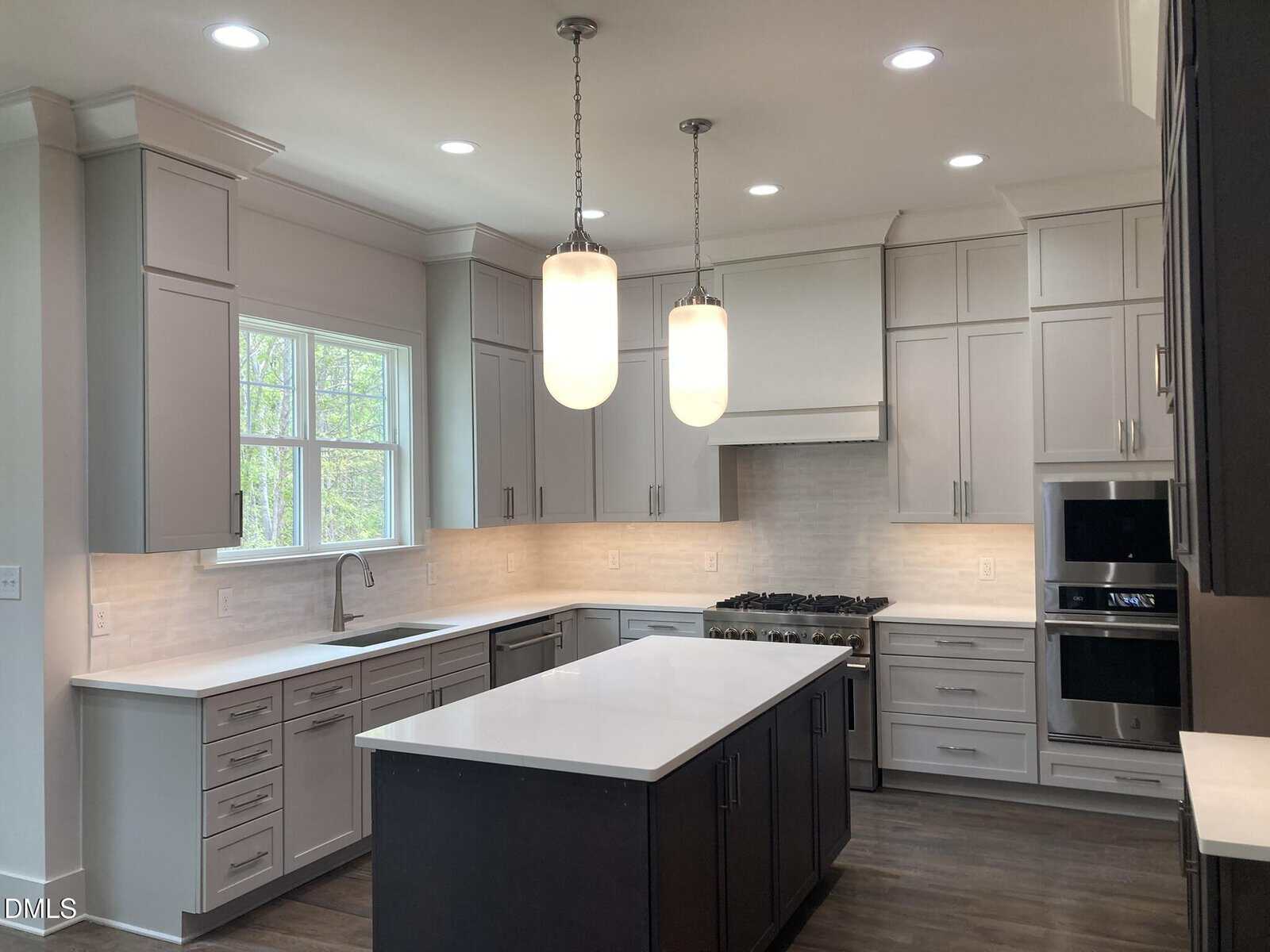 Modern kitchen with white cabinets, a large central island with dark base, stainless steel appliances, two pendant lights, and a window over the sink. Light countertops and backsplash create a bright, clean look.