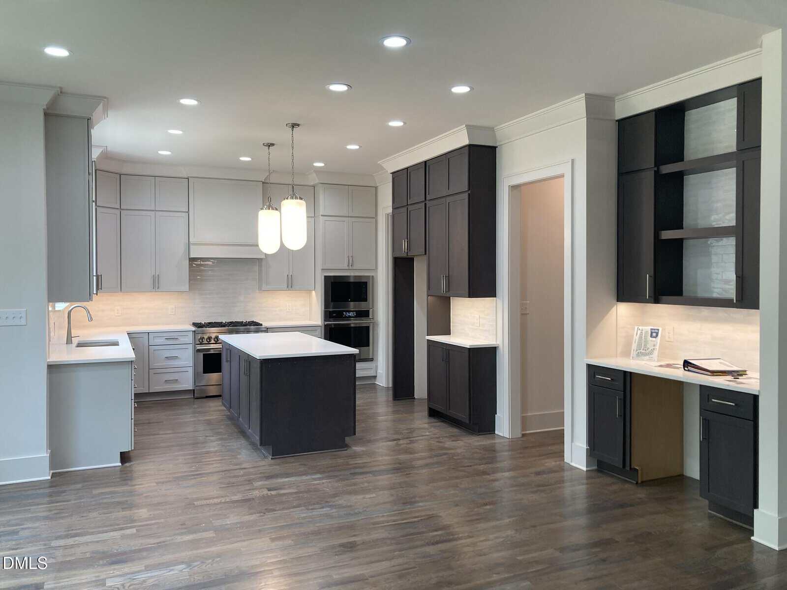 Modern kitchen with dark and light cabinets, a central island, built-in appliances, pendant lighting, a small desk area, hardwood floors, and recessed ceiling lights. The space feels open and contemporary.