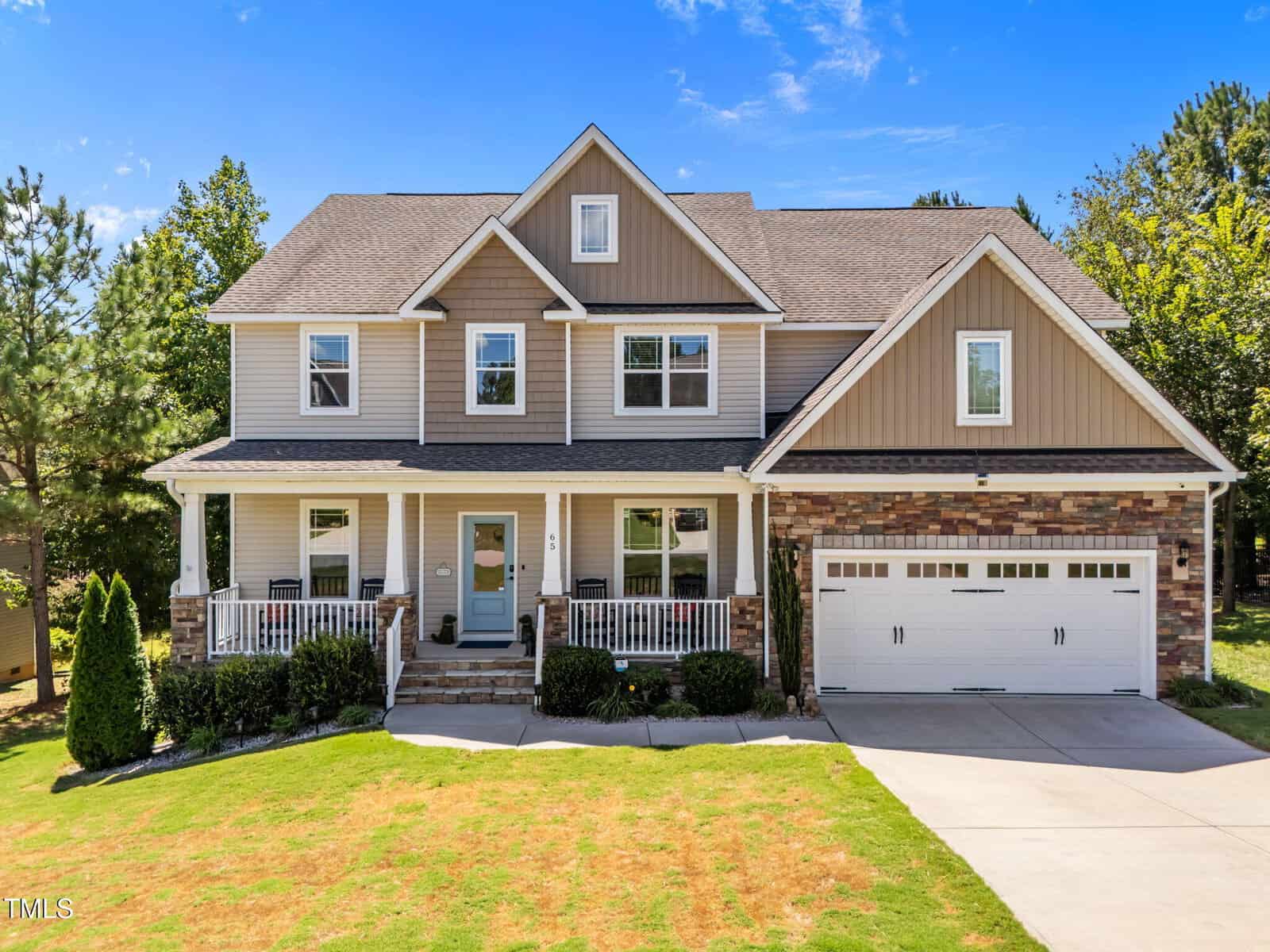 Two-story suburban house with tan siding, stone accents, a covered front porch, white railings, and a two-car garage. The front yard has green grass, bushes, and a concrete driveway under a clear blue sky.