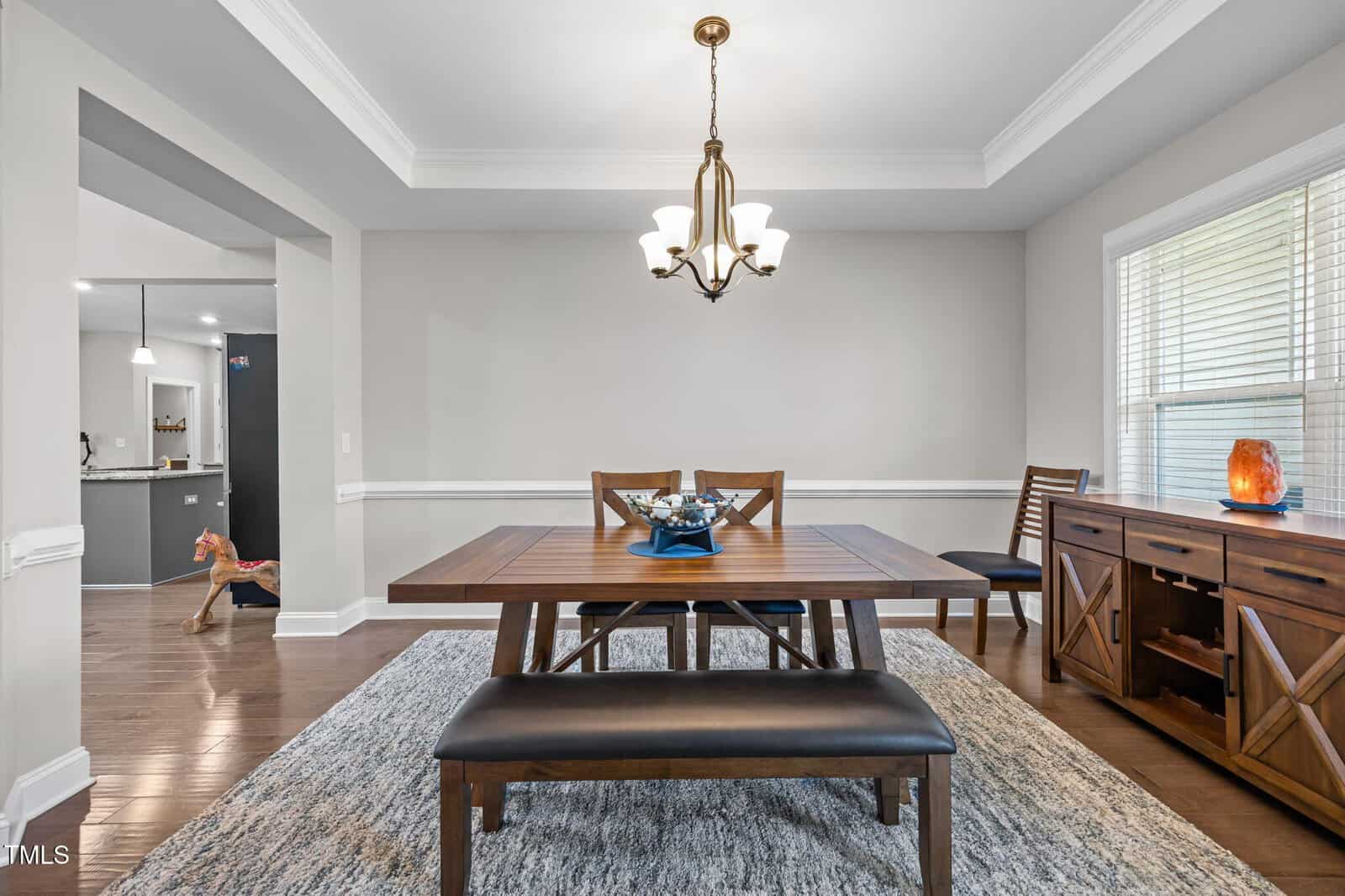 A dining room with a wooden table, two benches, a rug, a sideboard with a lamp, and a chandelier. A rocking horse is visible in an adjacent room. The walls are light gray, and windows let in natural light.