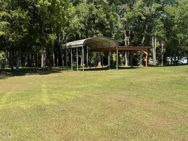 A metal carport and a wooden shelter stand on a grassy field bordered by tall, leafy trees under a clear sky.