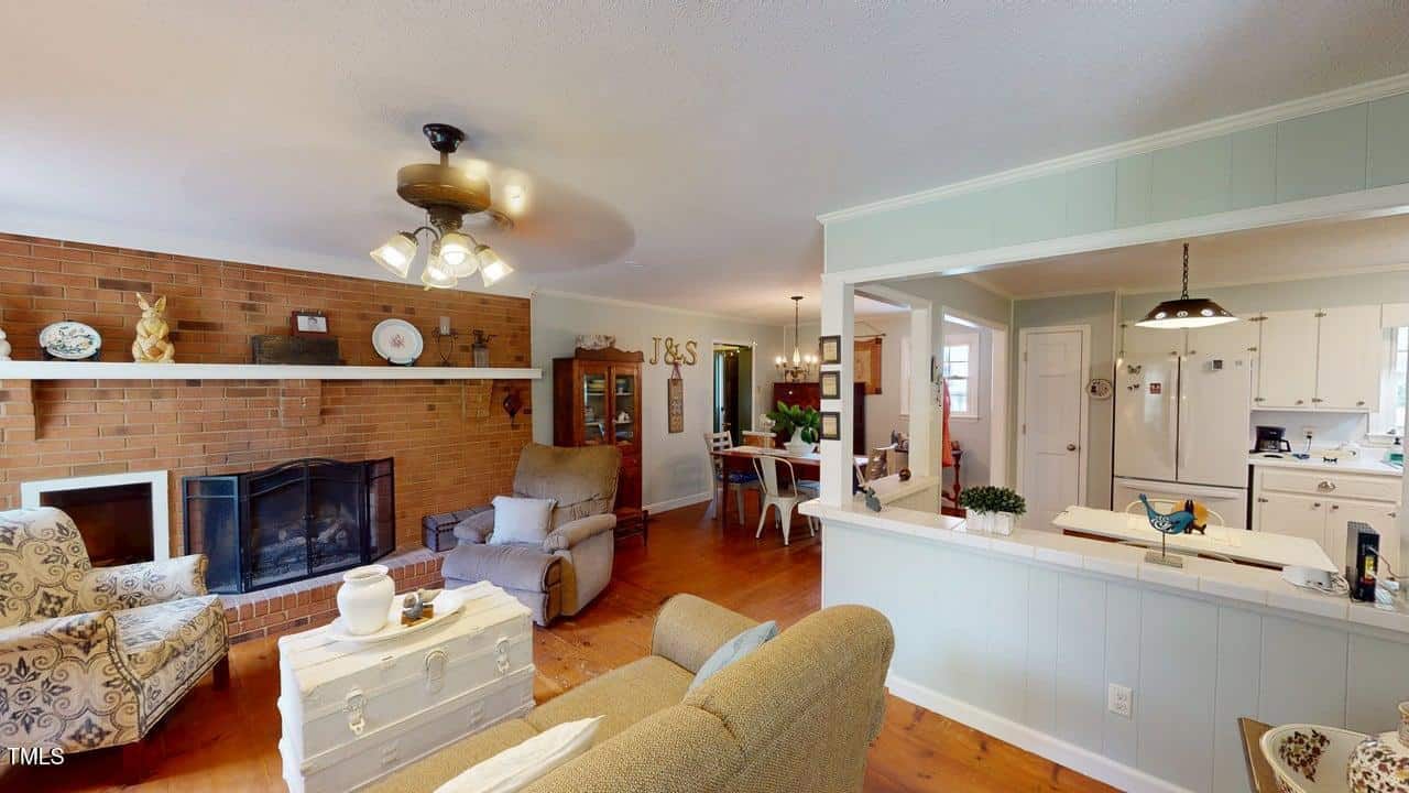 A cozy living room with a brick fireplace, armchairs, and a chest-style coffee table; an open layout leads to a dining area and a white kitchen, all with wood flooring and light-colored walls.
