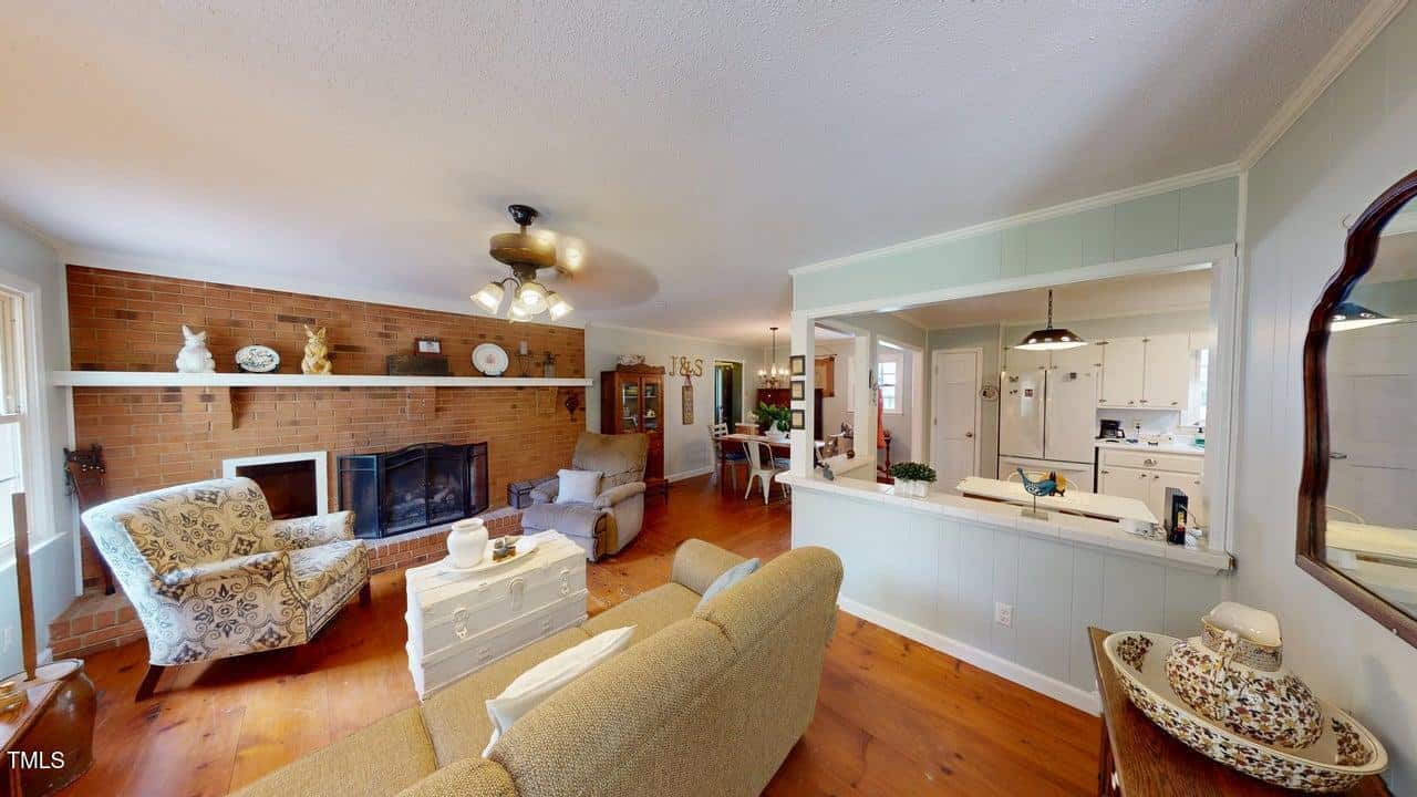 A living room with wood floors, a brick fireplace, armchairs, a sofa, and decorative items. An open window-style cutout in the wall leads to a white kitchen. Ceiling fan with lights is on the ceiling.