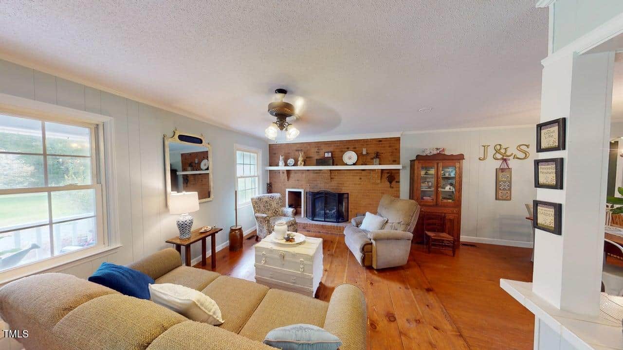 A cozy living room with hardwood floors, a brick fireplace, beige and blue furniture, a ceiling fan, a large window, wooden cabinets, and decorative items including "J&S" wall letters.