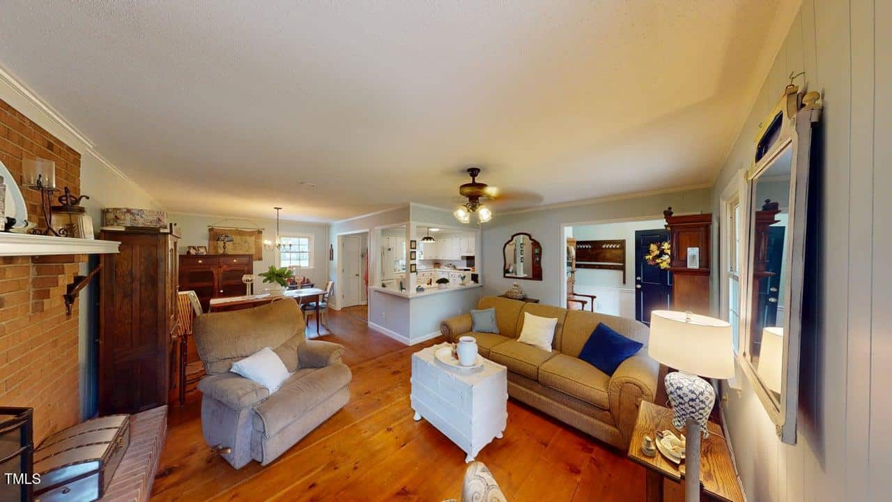 A cozy living room with wooden floors, a beige sofa, an armchair, a white coffee table, and a fireplace. The space opens to a dining area and kitchen, with decor in neutral and earthy tones.