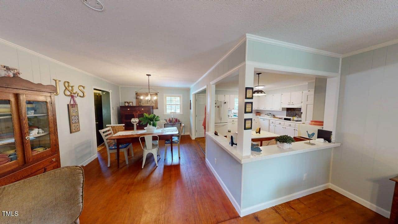 A dining area with a rectangular table and chairs is adjacent to a kitchen with white cabinets and countertops, separated by a half-wall. The space has wood floors and light-colored walls with decorative items on display.