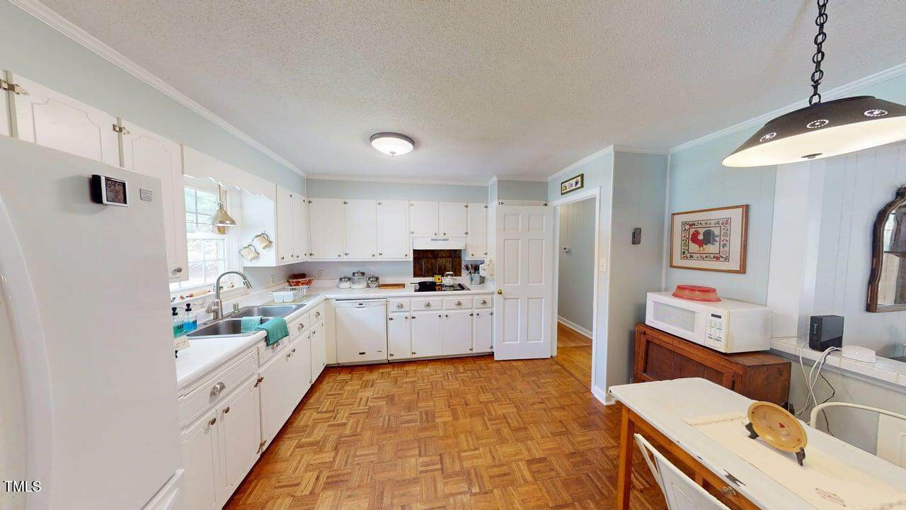 A bright kitchen with white cabinets, a farmhouse sink, wooden floor, and a small dining area with a table, chairs, microwave, and wall art. Natural light comes in through the window above the sink.