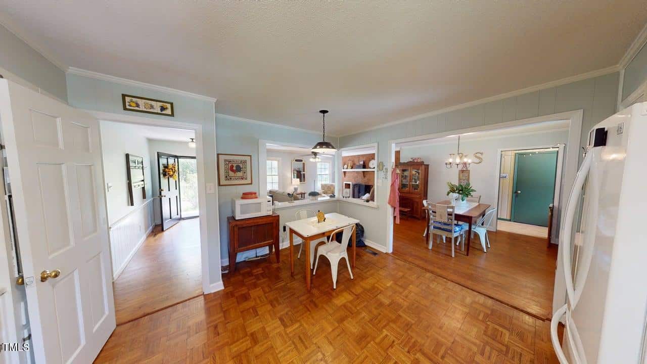 A cozy kitchen with a small dining table, white chairs, hardwood floors, and light blue walls opens to a dining room and hallway. A microwave sits on a side table, and a fridge is visible on the right.
