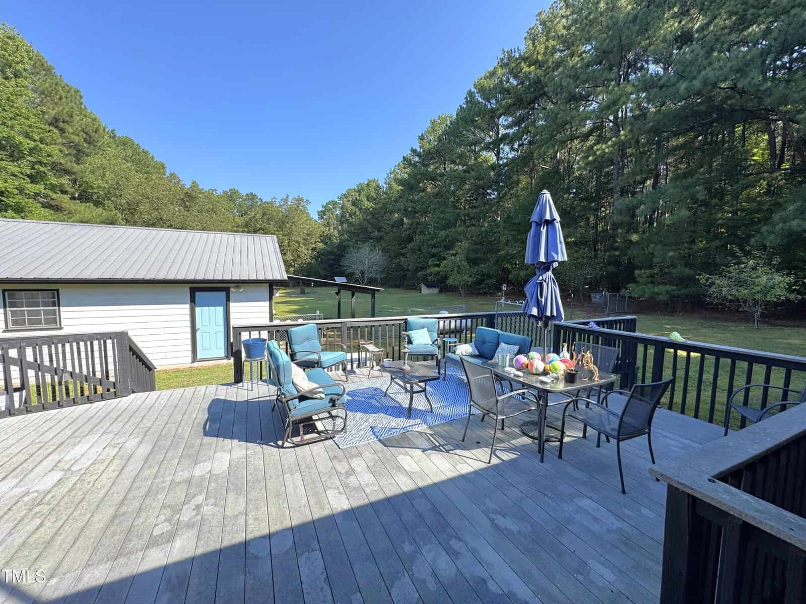 A large wooden deck with outdoor furniture, including cushioned chairs, tables set with drinks and flowers, and a blue umbrella. Trees and a white outbuilding with a blue door are visible in the background.