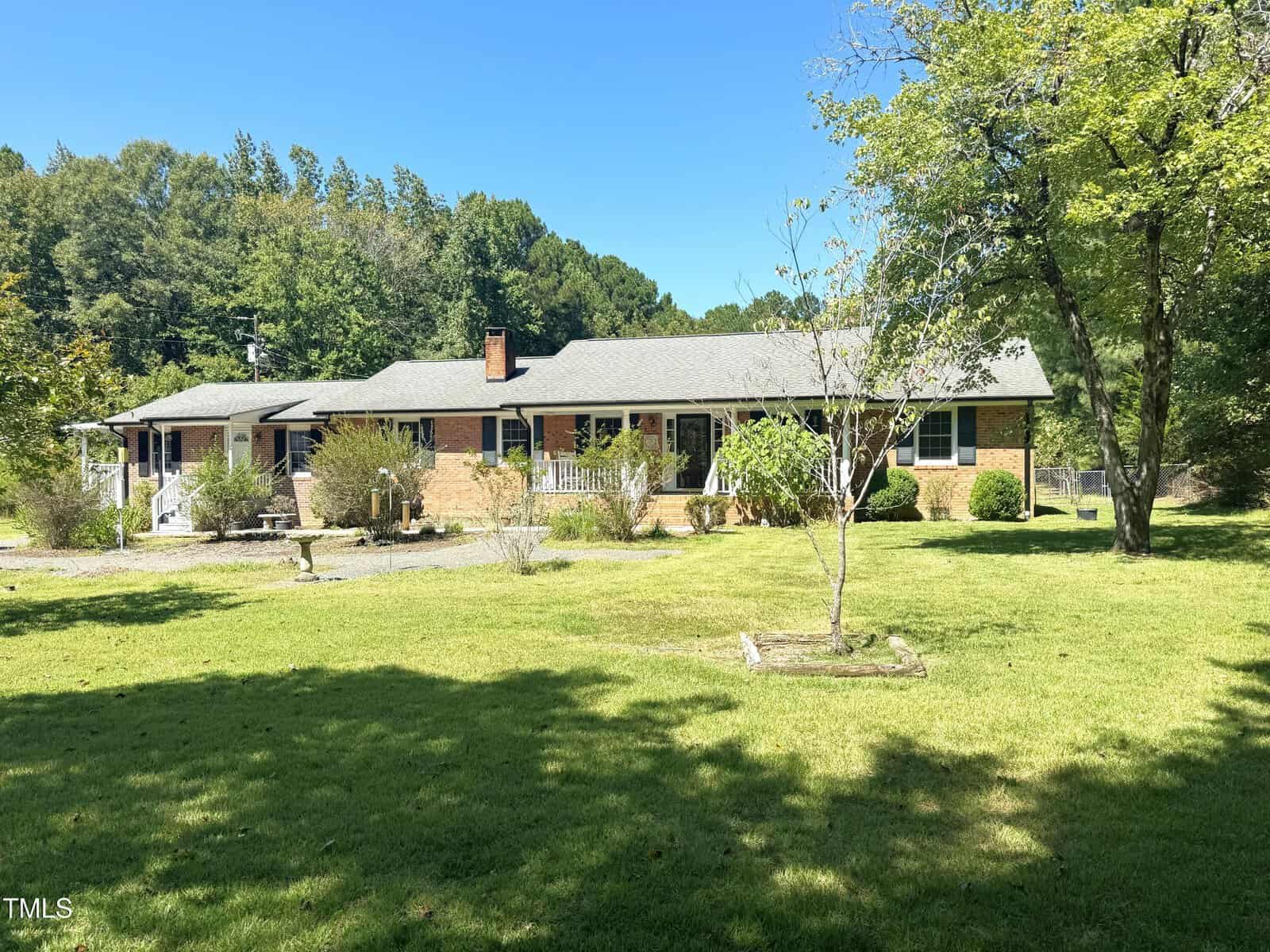 A single-story brick house with a gray roof sits on a large grassy lawn at 1079 Fate Washington Road in Stem. Surrounded by trees and shrubs, the yard features a small birdbath, a gravel driveway, and clear blue skies above.