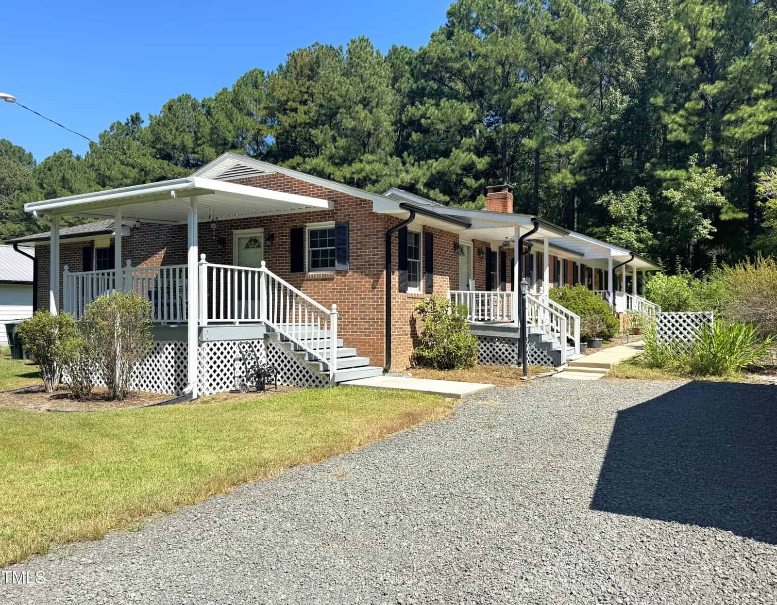 A one-story brick house with a covered front porch, white railings, and black shutters sits beside a gravel driveway. The yard has bushes, lawn, and is bordered by tall trees under a clear blue sky.