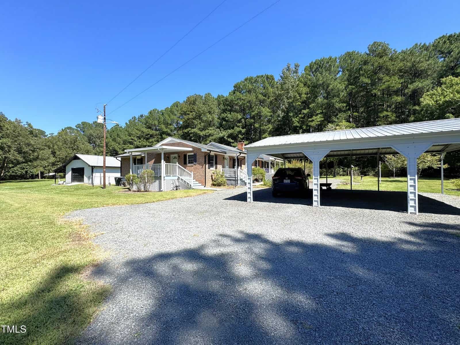 A single-story brick house with a front porch is set in a grassy yard with trees in the background. A gravel driveway leads to a covered carport with vehicles parked underneath. A small outbuilding is visible to the left.
