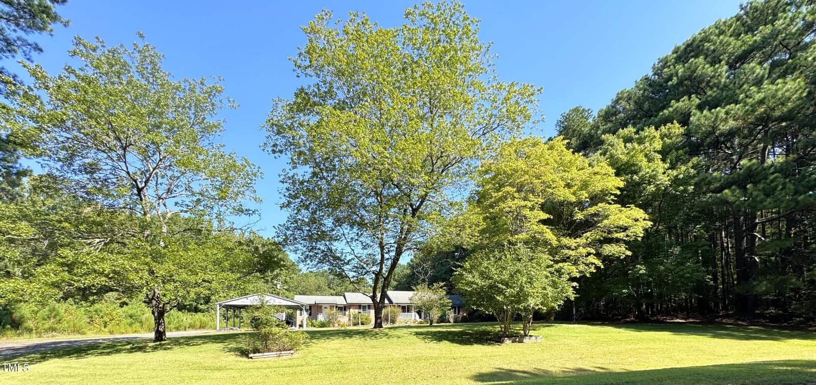 A single-story house with a covered carport sits in the background, surrounded by large green trees and a well-maintained grassy yard under a clear blue sky.