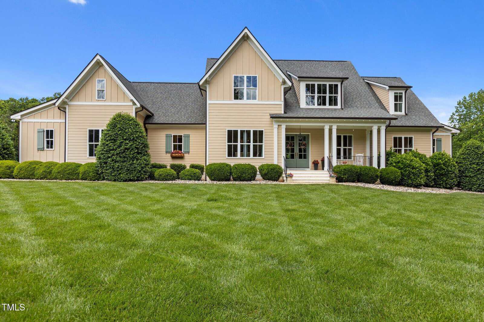 A large beige house with dark gray roof, white trim, multiple gables, and a covered front porch. The house is surrounded by neatly trimmed bushes and a well-maintained green lawn under a clear sky.