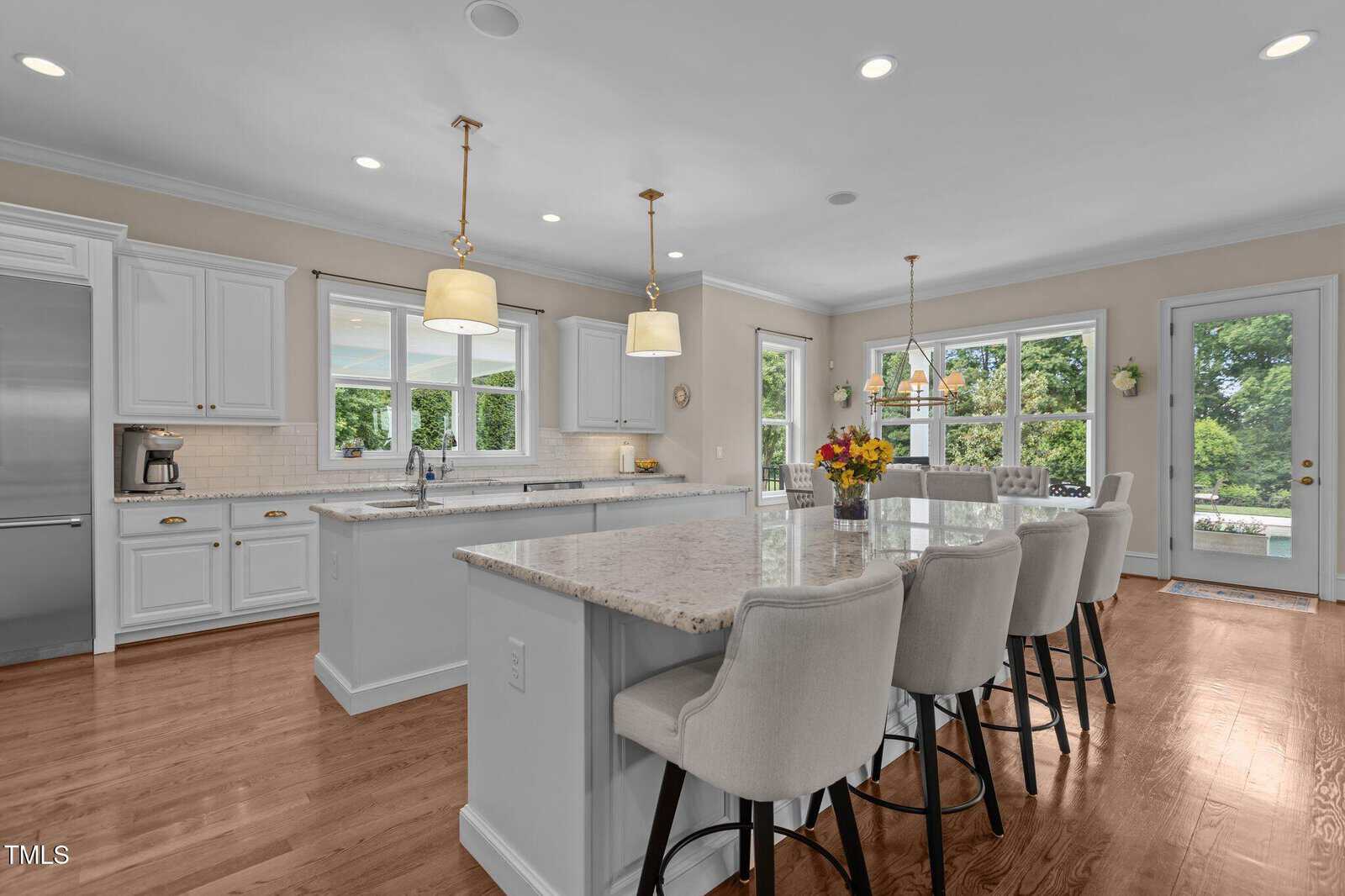 Spacious modern kitchen with white cabinets, marble countertops, large island with bar stools, pendant lights, wood floors, and windows overlooking a green backyard with a pool.