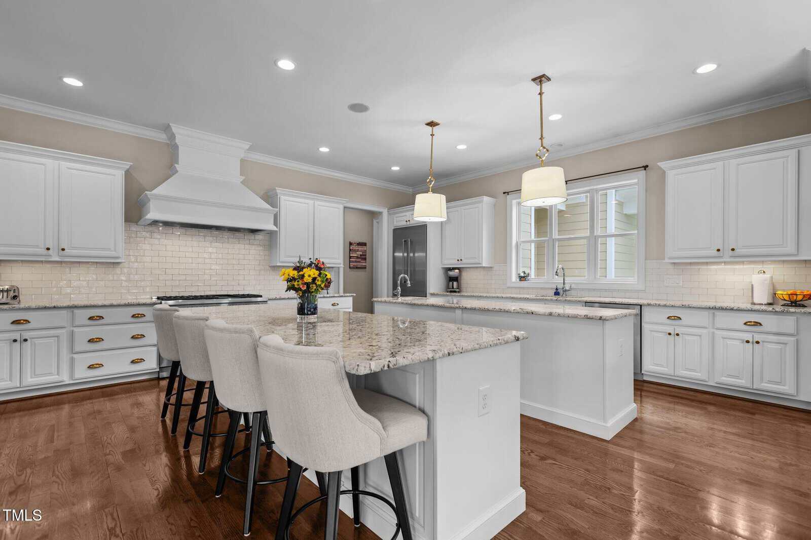 Modern kitchen with white cabinets, marble countertops, stainless steel appliances, a large island with four upholstered barstools, pendant lights, hardwood floors, and a vase of sunflowers on the island.