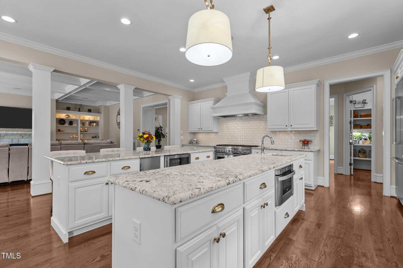 Spacious kitchen with white cabinets, marble countertops, hardwood floors, stainless steel appliances, two islands, pendant lights, and an open view to the living room and pantry.