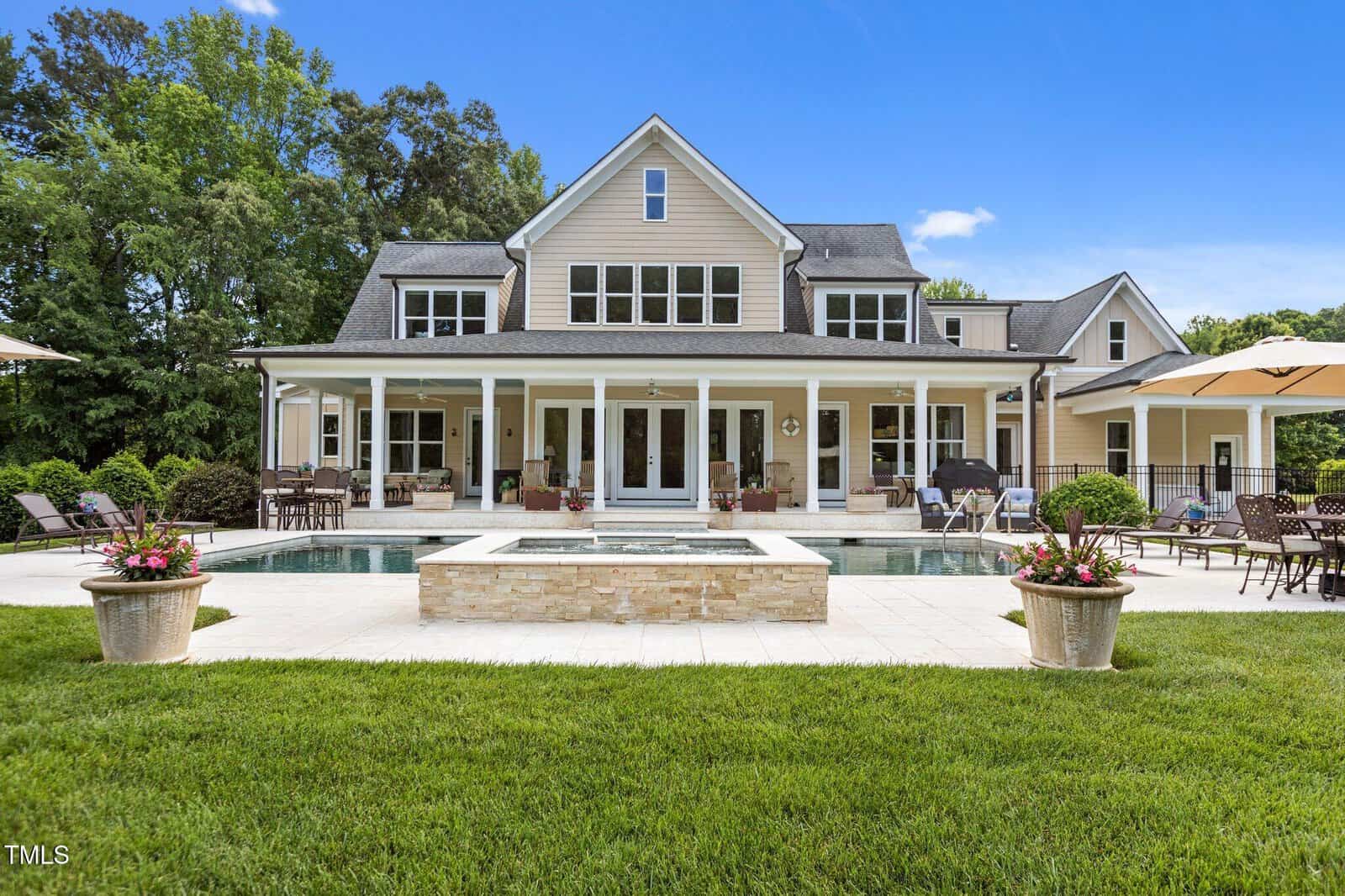 A large beige house with multiple gables and white trim features a covered porch, outdoor seating, a rectangular pool with a stone edge, and planters with flowers, all set against green grass and trees under a blue sky.
