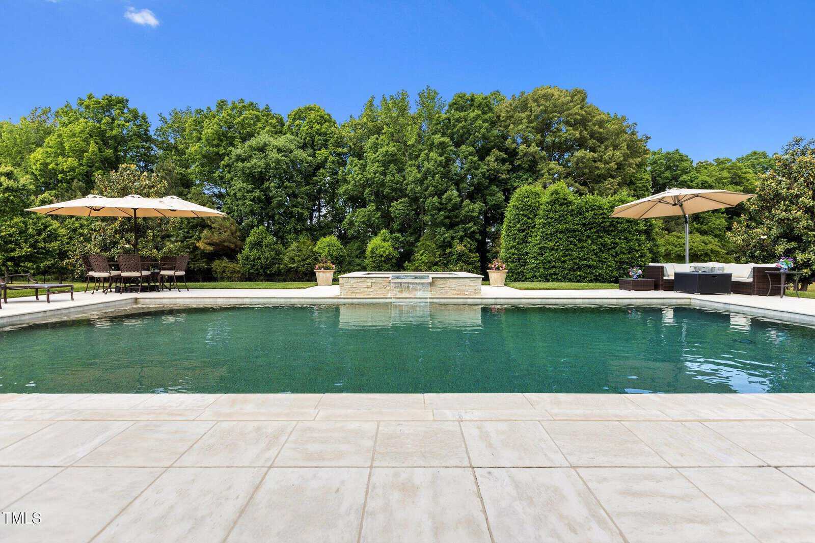 A rectangular swimming pool with clear water, surrounded by a stone patio. There are lounge chairs, tables, and large beige umbrellas on both sides. Green trees and bushes border the background under a clear blue sky.