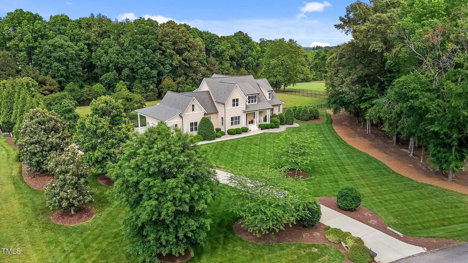 A large two-story house with a gray roof and light-colored exterior is surrounded by lush green lawns, trees, and landscaped shrubs, set against a backdrop of dense forest under a blue sky.