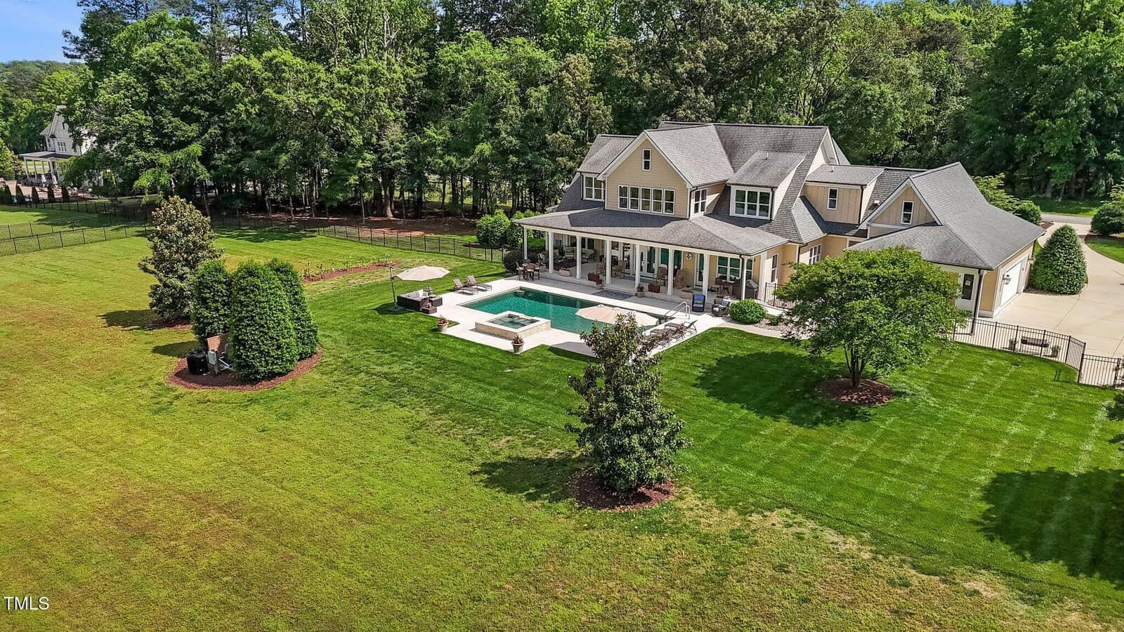 A large house with a gray roof and beige exterior sits on a spacious green lawn, featuring a backyard pool, hot tub, patio seating, and surrounded by trees and landscaping.