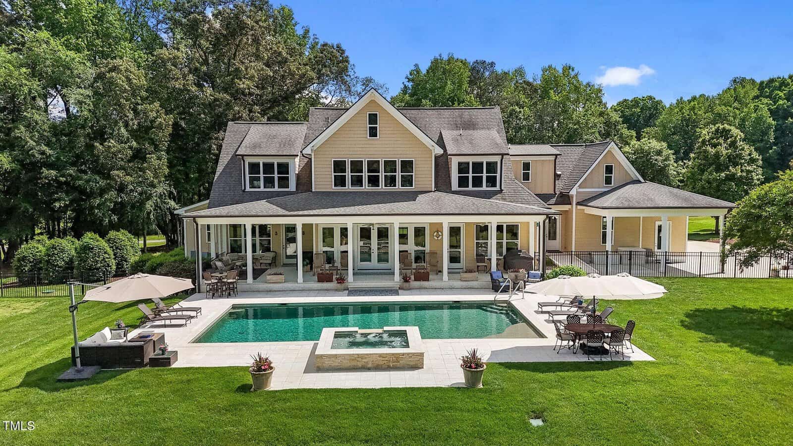 A large, beige two-story house with dormer windows, a covered back porch, and an in-ground rectangular pool with a hot tub, surrounded by green lawn, trees, and outdoor seating areas.