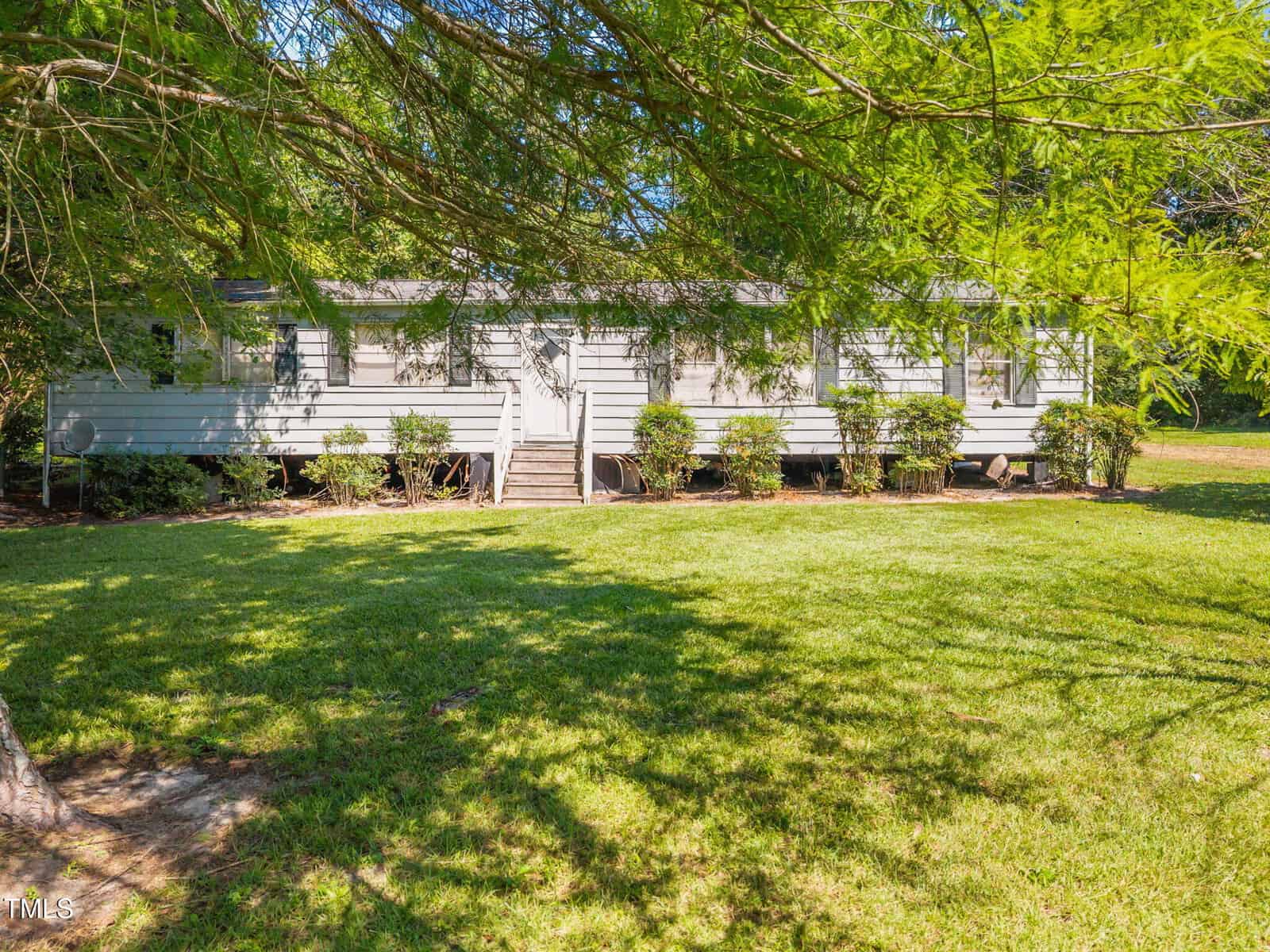 A single-story white manufactured home with a small front stoop, green shrubs along the front, and a well-maintained grassy yard with large trees providing shade.