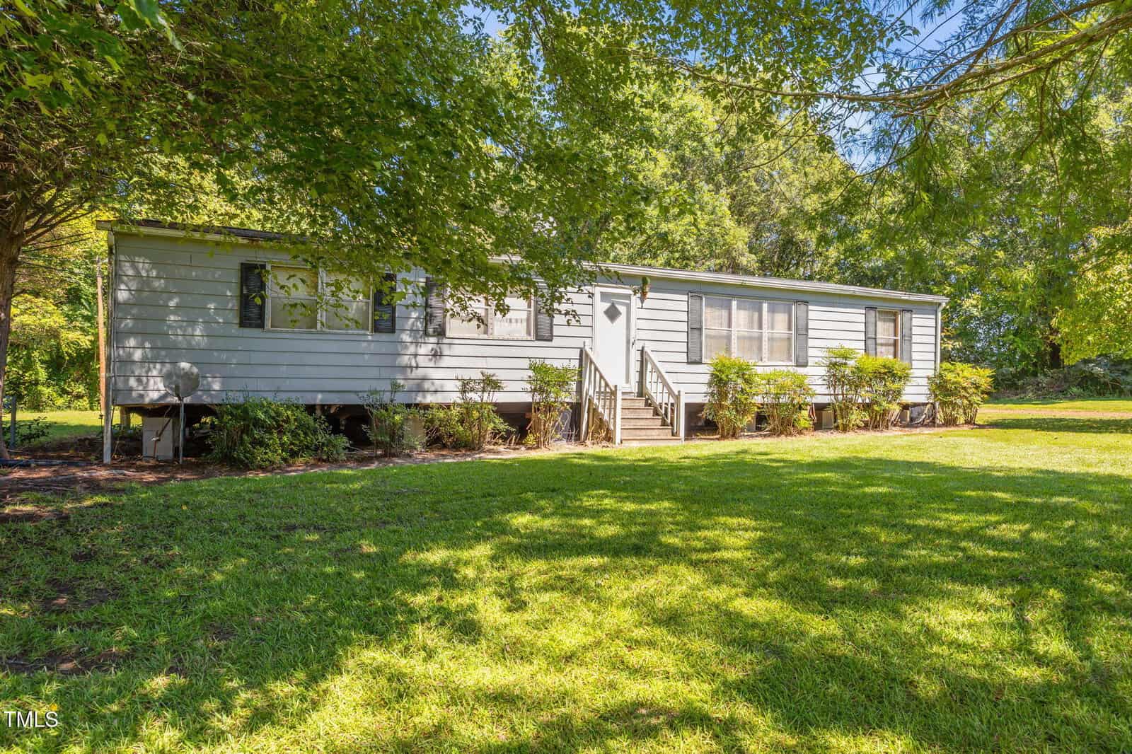A single-story, light gray manufactured home with several windows and a small front stoop, sits on a grassy lawn surrounded by trees and shrubs in bright daylight.