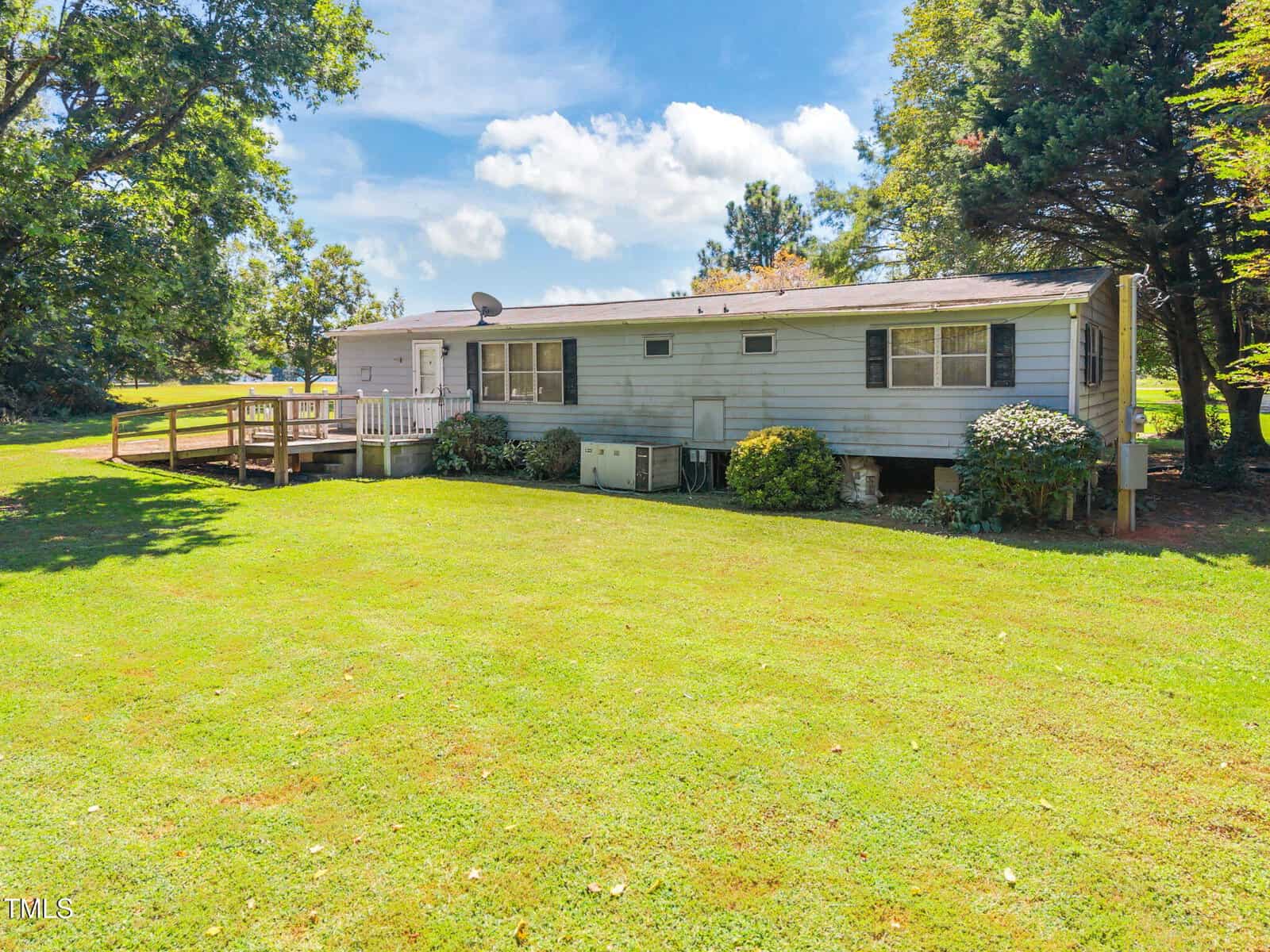A single-story light blue manufactured home with black shutters, a wooden ramp, and small bushes in front, situated on a large grassy yard surrounded by mature trees under a partly cloudy sky.