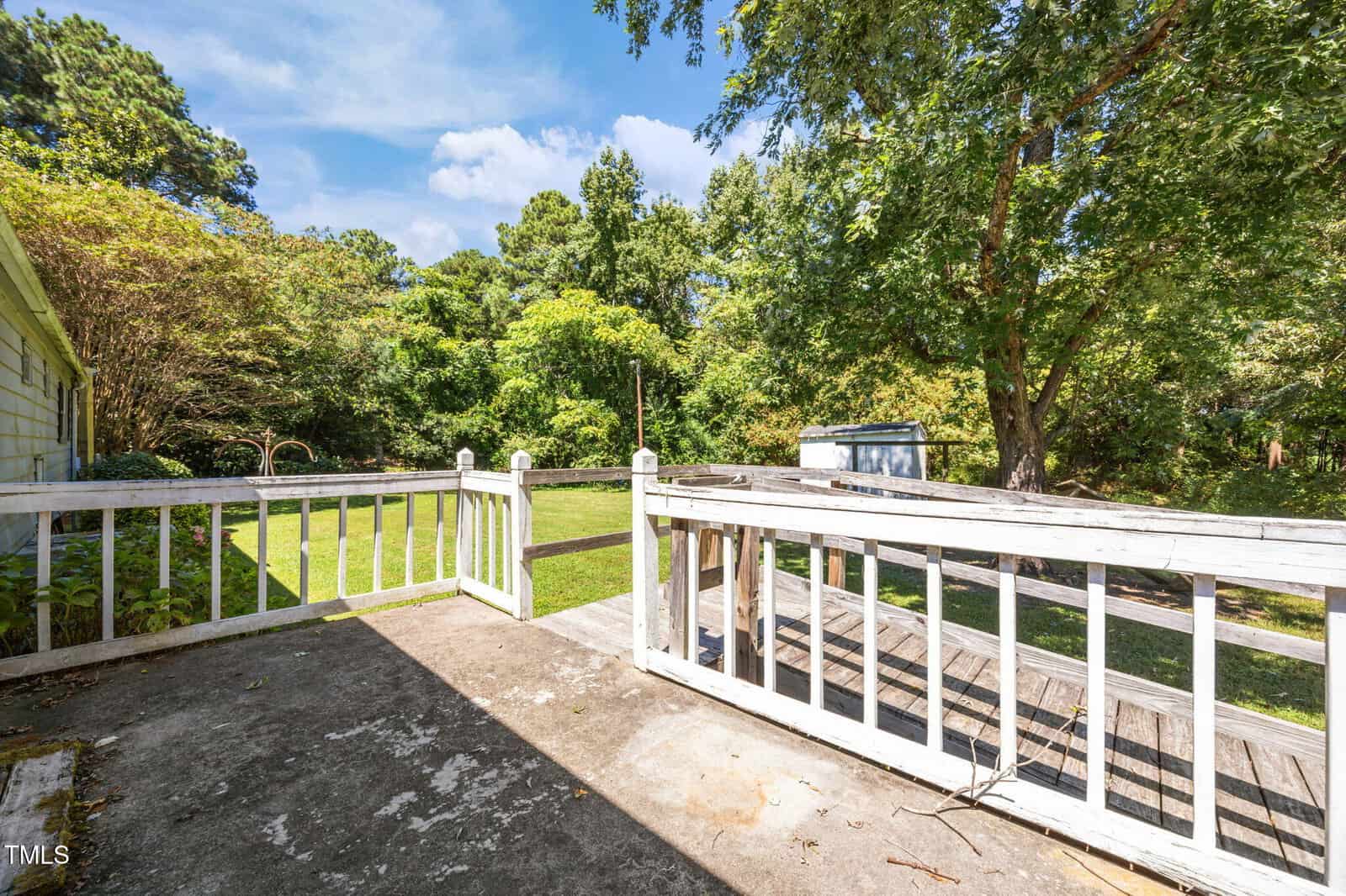 A view from a wooden deck with white railings looking out onto a grassy backyard with trees, a shed, and a blue sky with clouds. The deck has some worn areas and overgrown plants nearby.