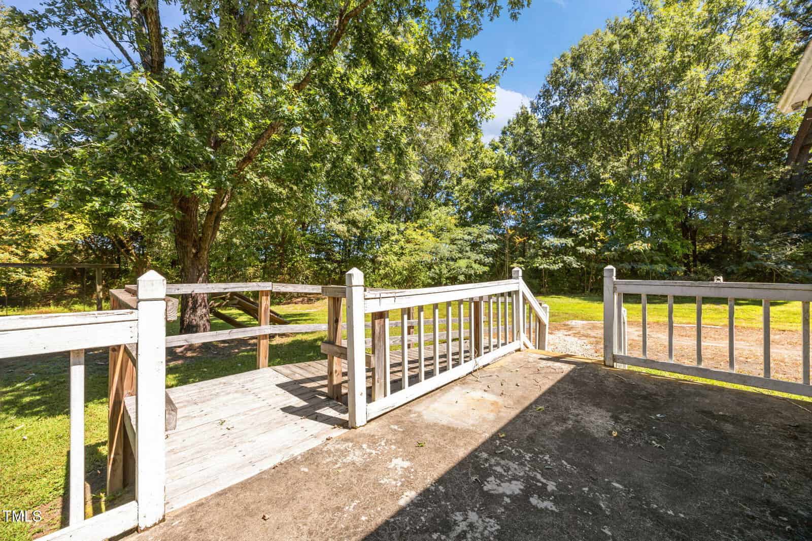 A wooden ramp with white railings leads from a concrete patio to a grassy yard surrounded by trees under a clear blue sky.