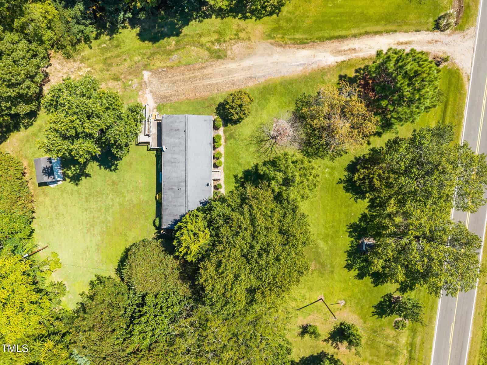 Aerial view of a house surrounded by trees and a grassy lawn, with a driveway leading to a road on the right side. Shadows from trees are visible on the grass.