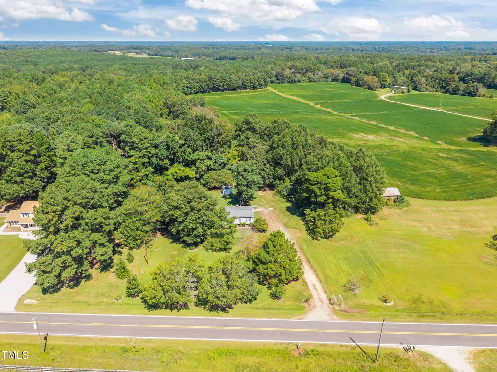 A rural property with a house surrounded by trees, a driveway, open grassy fields, and nearby farmland, with a road in the foreground and a wooded area in the distance under a partly cloudy sky.