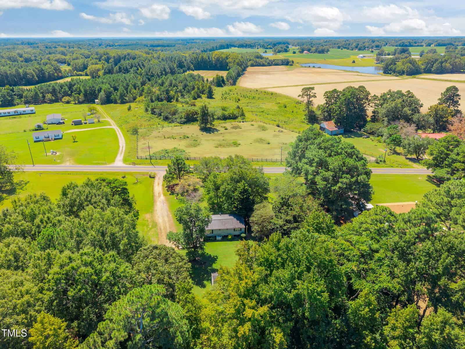 Aerial view of a rural landscape with open fields, scattered trees, a few houses, and a winding road under a partly cloudy sky.