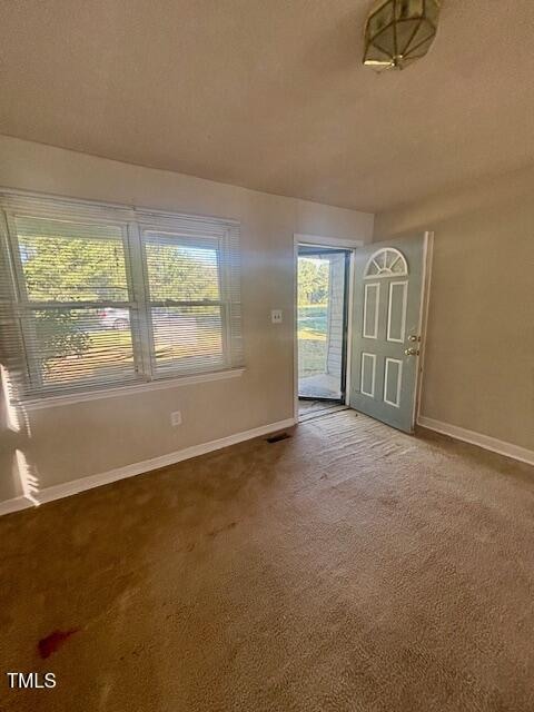 A small room with beige carpet and light-colored walls, featuring a window with blinds on the left and a front door partially open to the outside. Sunlight is coming through the window.