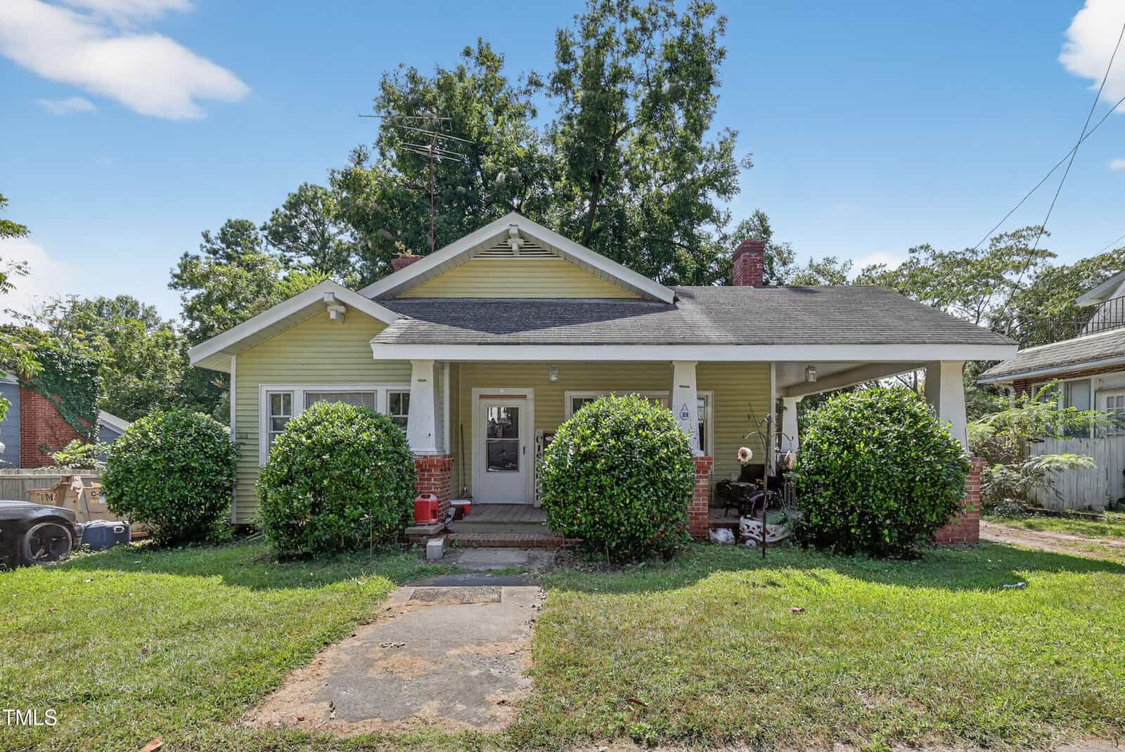 Single-story house with yellow siding, a covered front porch, and white trim. Two large bushes frame the entrance, and a tree stands behind the roof. The lawn is green, with a sidewalk leading to the front door.